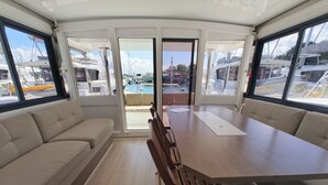 A boat interior with a dining area, cushioned seating, and large windows offering views of the marina.