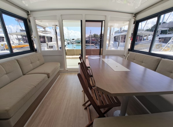 Interior de un barco con comedor, asientos acolchados y ventanas amplias con vistas a otros barcos y un puerto deportivo.