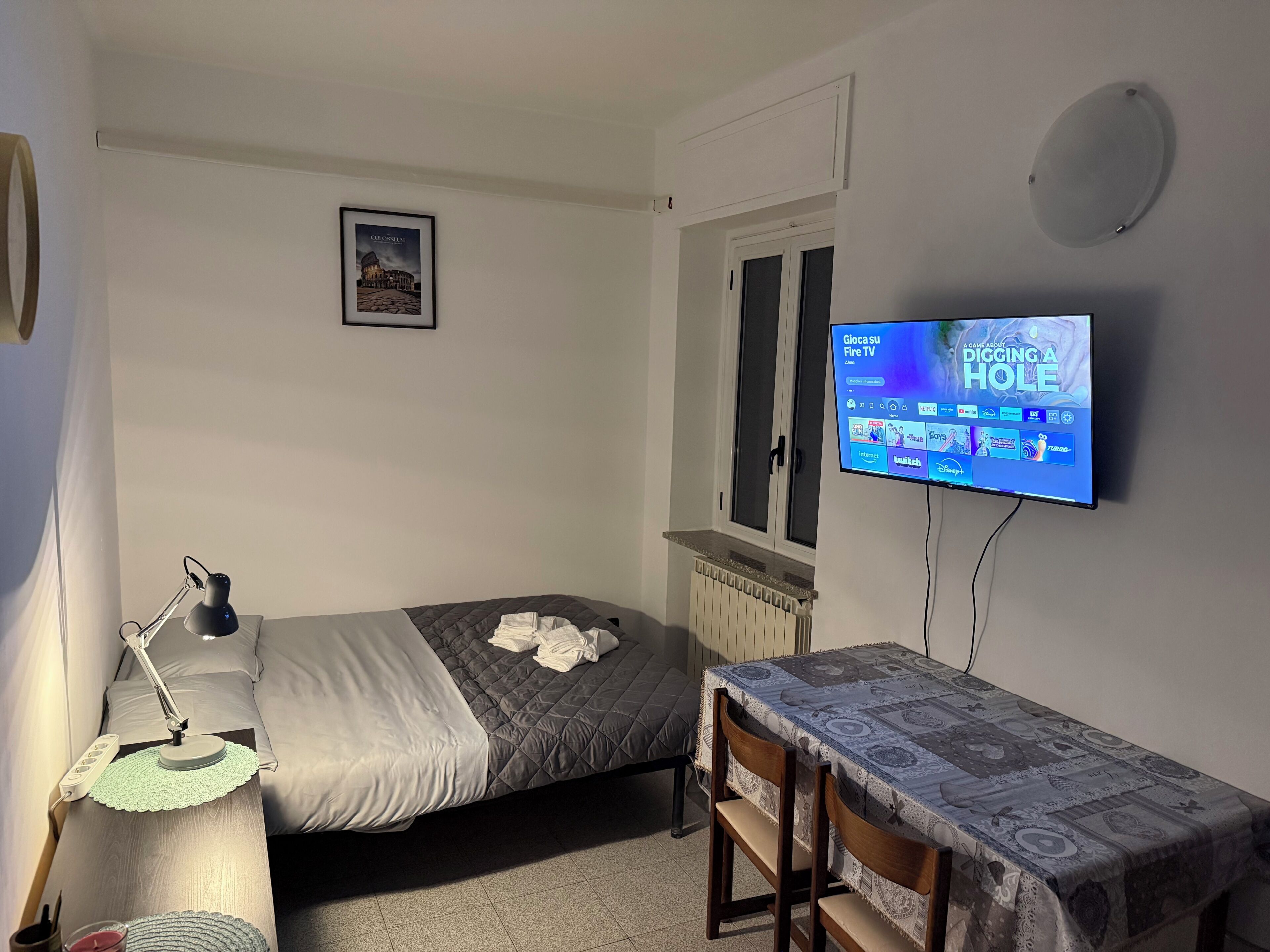 A bedroom with a bed, bedside table, lamp, TV, and a window.