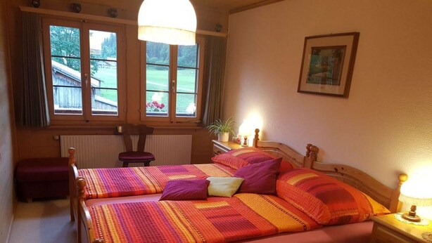 A bedroom with a bed, a chair, a bedside table, a lamp, and a window with curtains. - Steimandli Appartement/Fewo, Dusche oder Bad, WC 2-Bettwohnung by Interhome (Gstaad)
