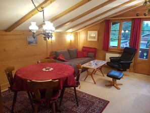 Ein gemütliches Zimmer mit Holzvertäfelung, einem Esstisch mit Stühlen, einem Sofa, einem Sessel und einem Fenster mit Vorhängen.