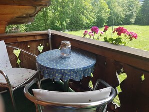 Eine Holzterrasse mit einem runden Tisch und Stühlen, einem Metallstuhl und Blick auf eine grüne Wiese mit rosa Blumen.