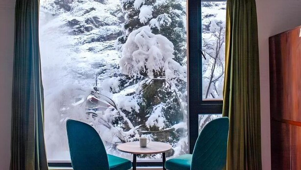 A room with a large window, two teal chairs, a small round table, and a cup. - Mystique Valley by Trekocity  (Lachung)