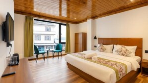 A hotel room with a wooden ceiling, a bed with white bedding, a TV mounted on the wall, a small table with a chair, and a window with curtains. - Mystique Valley by Trekocity  (Lachung)