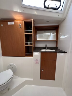 A compact bathroom with a white toilet, a sink, and a mirror above it. There is a wooden cabinet with a shelf and a small plant. - Sun-Kissed Sailing Adventure in Zadar, Croatia (Bibinje)