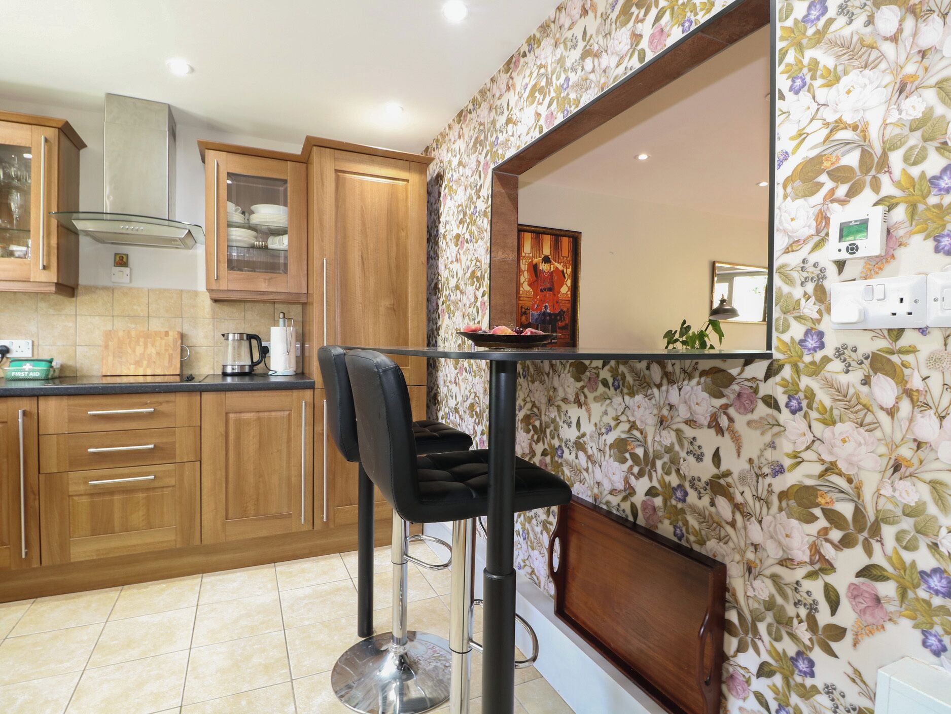 A modern kitchen with wooden cabinets, a stainless steel range hood, a bar stool, and a floral wallpaper.