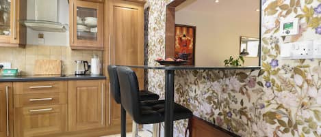 A modern kitchen with wooden cabinets, a stainless steel range hood, a bar stool, and a floral wallpaper.