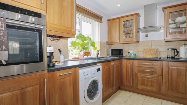 Een keuken met houten kastjes, een ingebouwde oven, een magnetron, een wasmachine en een raam met een plant.