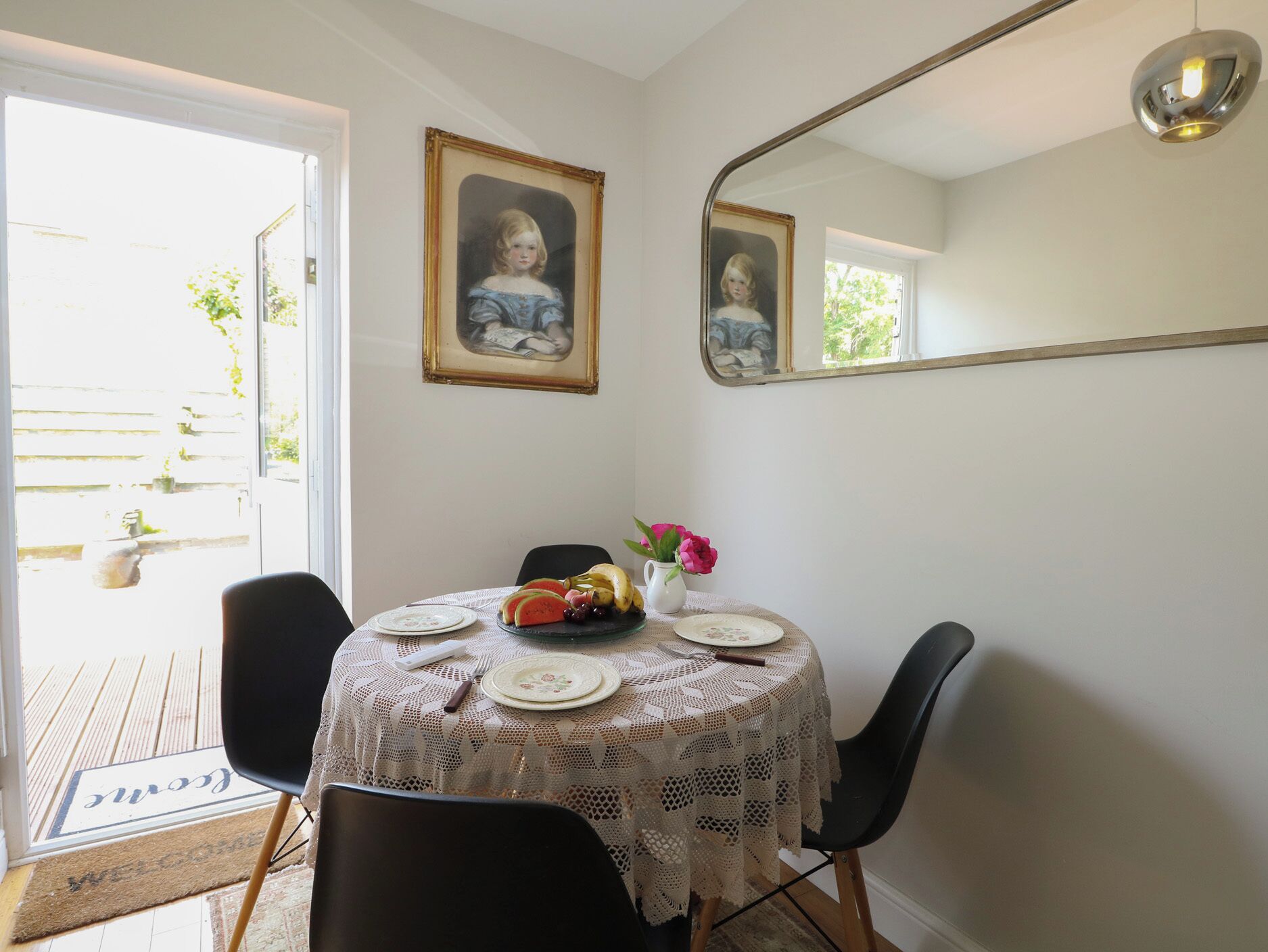 A dining area with a round table, chairs, a lace tablecloth, a vase with flowers, and a framed portrait on the wall.