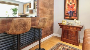 Een moderne keuken met een bar, twee barkrukken, een houten kastje met decoratieve spullen, en een grote spiegel die de keuken weerspiegelt.