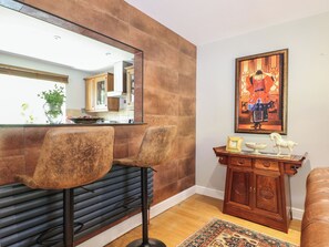 A modern kitchen with a bar area, two bar stools, a wooden cabinet with decorative items, and a large mirror reflecting the kitchen. - The Dacha (Winchester)