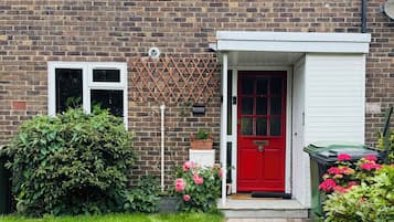 A brick house with a red door, a white window, and a small garden with pink flowers.