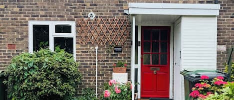 Una casa de ladrillo con puerta roja, ventana blanca y un pequeño jardín con flores rosadas.
