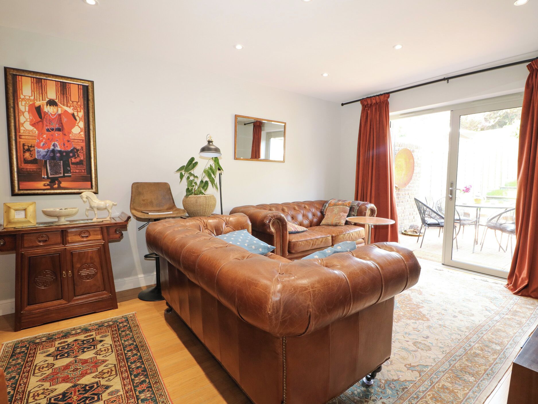 Un salon avec un canapé en cuir marron, un meuble en bois, une lampe sur pied, une plante en pot et une grande fenêtre avec des rideaux.
