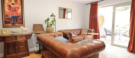 Un salon avec un canapé en cuir marron, un meuble en bois, une lampe sur pied, une plante en pot et une grande fenêtre avec des rideaux.