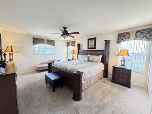 Quarto com cama, mesa de cabeceira, cômoda, um banquinho, ventilador de teto e janela com persianas.