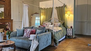 A bedroom with a four-poster bed, a sitting area with a chair and table, and a nightstand with a lamp.
