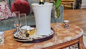 A room with a round table featuring a white vase with a gold candle holder, a glass tray with a gold bowl, a red rose in a glass vase, and a patterned sofa with a red cushion.