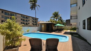A swimming pool with two wicker chairs, a small covered seating area, and palm trees. - Sea Residence (Ipojuca)