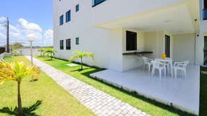 A modern building with a flat roof, a covered patio with white chairs, and a small garden with palm trees. - Sea Residence (Ipojuca)