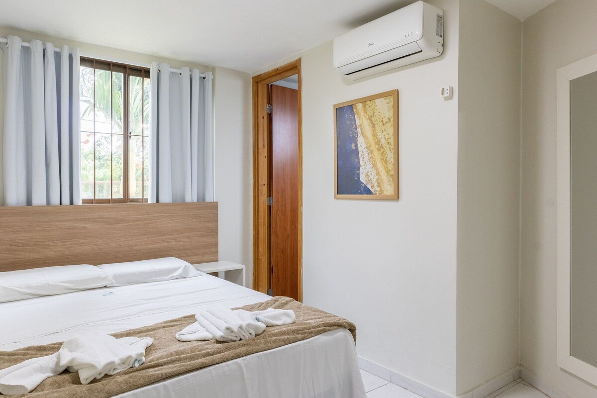 A hotel room with a bed, an air conditioner, a door, and a painting on the wall.