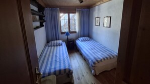 Ein kleiner, gemütlicher Raum mit zwei Einzelbetten, einem Fenster mit Vorhängen, einer Lampe und einer Holzbalkendecke.