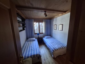 Ein kleiner, gemütlicher Raum mit zwei Einzelbetten, einem Fenster mit Vorhängen, einer Lampe und einer Holzbalkendecke.