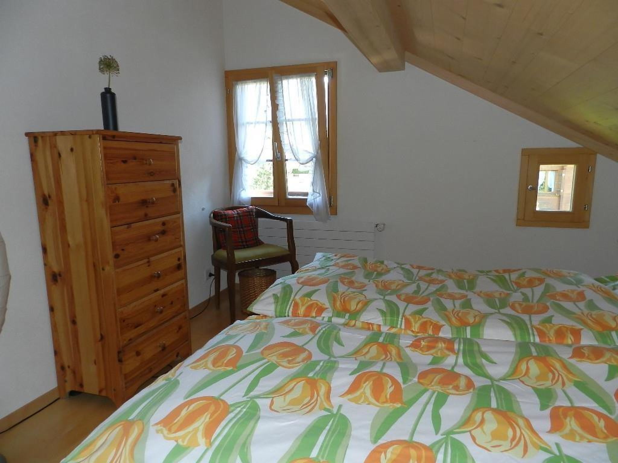A bedroom with a wooden dresser, a chair, a bed with a floral bedspread, a window with curtains, and a skylight.