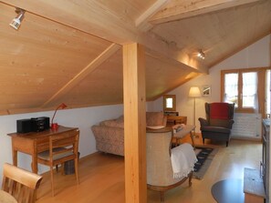 Een gezellige zolderkamer met houten balken, een bureau met elektronische apparatuur, een bank, een stoel en een eettafel.