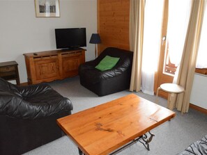 Ein Wohnzimmer mit Couchtisch aus Holz, einer schwarzen Ledersofa, einem Ledersessel mit grünem Kissen, einem Fernseher auf einer Holzkommode und einem kleinen Hocker.