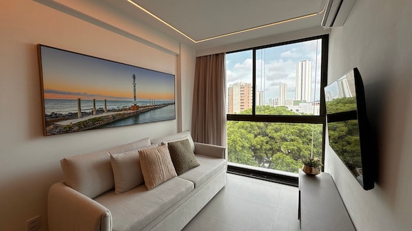 A modern living room with a large window, a sofa, a flat-screen TV, and a painting of a coastal scene.