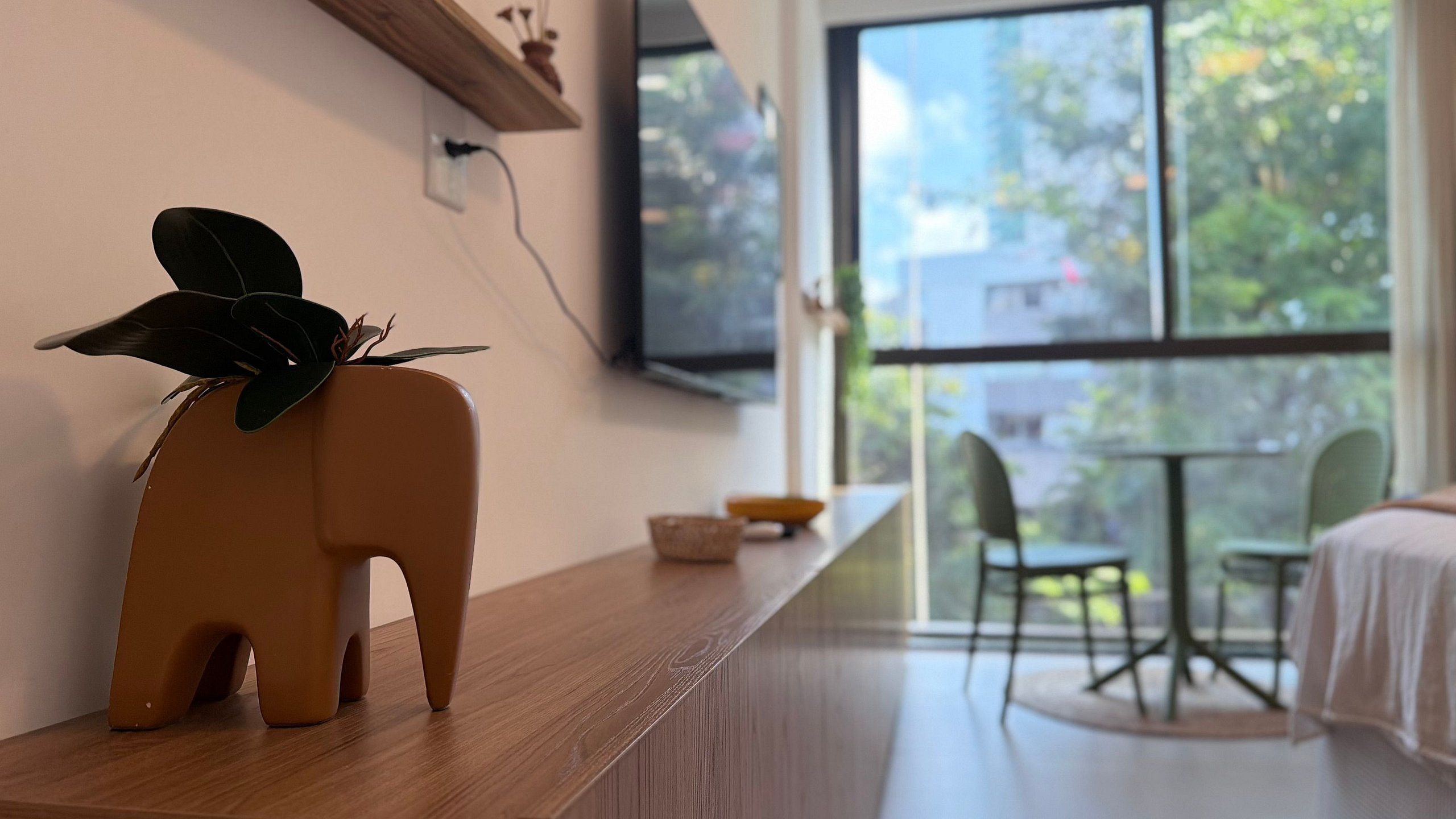 Quarto moderno com mesa de madeira, um suporte de planta em formato de elefante e vista para o verde através de janelas amplas.