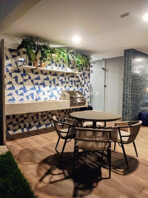 An outdoor patio with a round wooden table and chairs, a built-in grill, and a tiled backsplash with a geometric pattern. - Casa Forte Prince por Yolo (Recife)