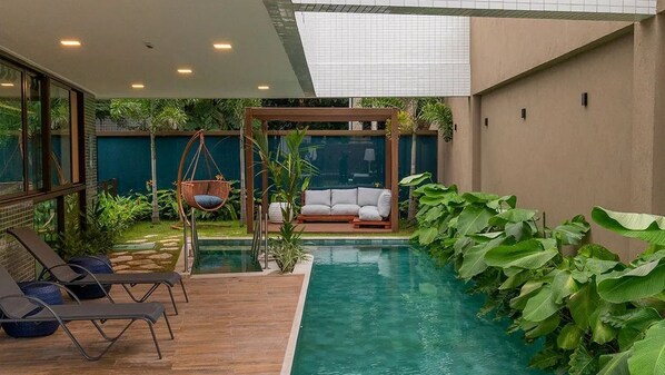 Un área de piscina moderna al aire libre con sillones de descanso, una hamaca colgante y una zona de estar con cojines.