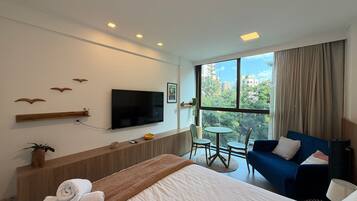 Una habitación de hotel con cama, televisor, una mesita con sillas, sofá y ventana con vista a edificios y árboles.
