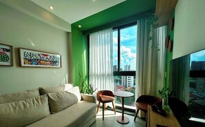 A modern living room with a sofa, a small round table, and chairs, a large window with curtains, and two framed artworks on the wall.