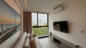 A modern living room with a large window, a flat-screen TV, and a painting of a coastal scene.