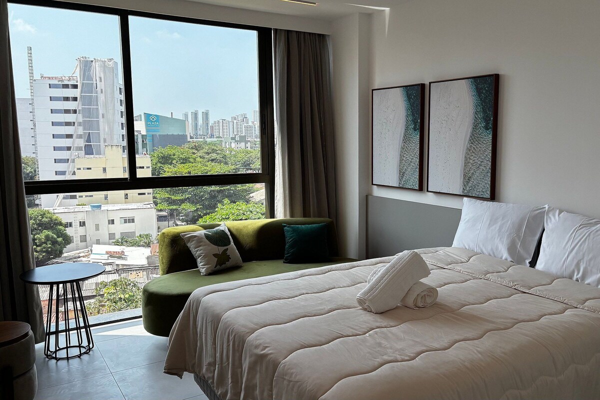 A hotel room with a large bed, a green sofa, a small table, and a view of the cityscape through large windows.