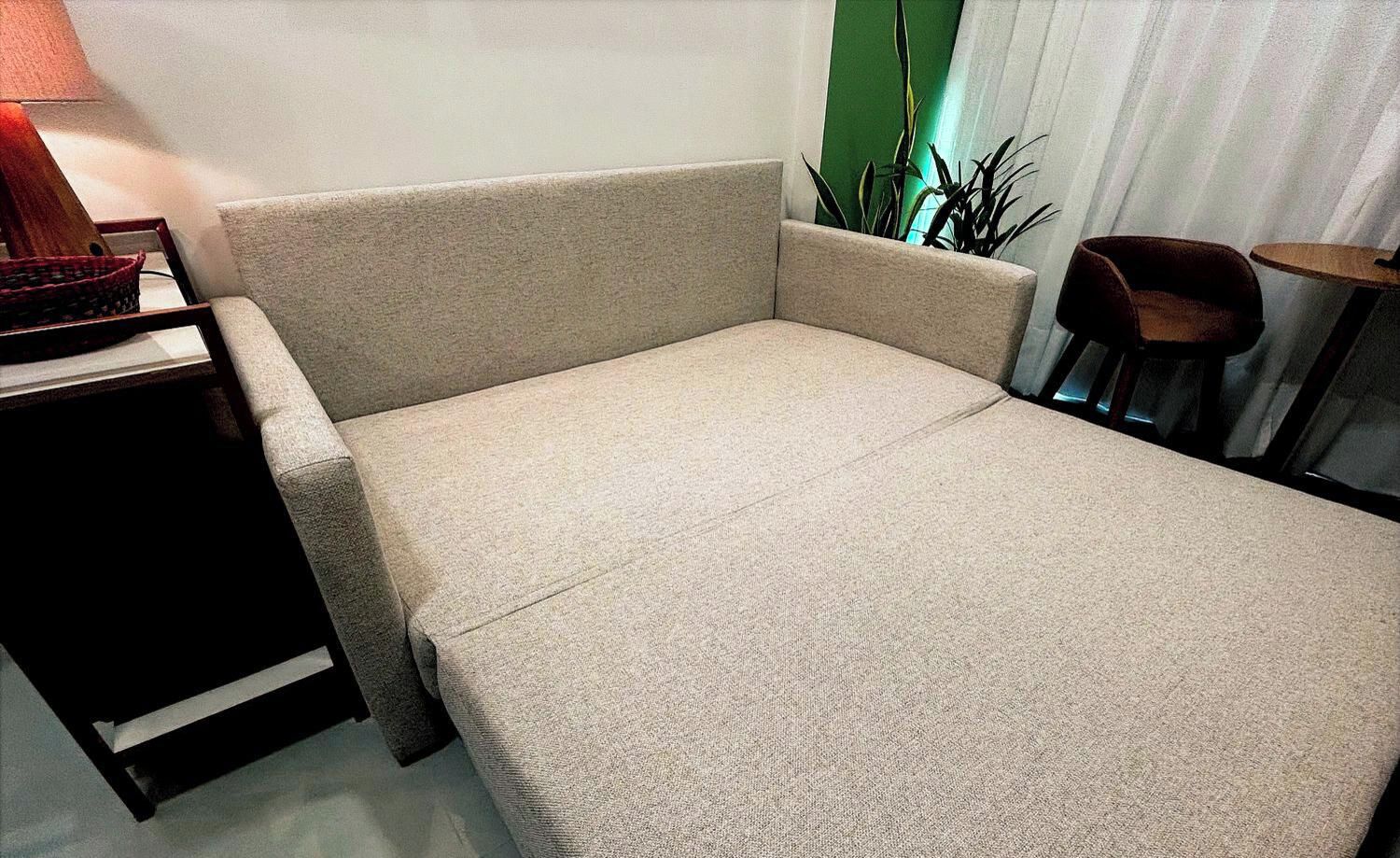 Quarto compacto com cama de solteiro, sofá-cama em tons neutros, mesa pequena e uma cadeira.
