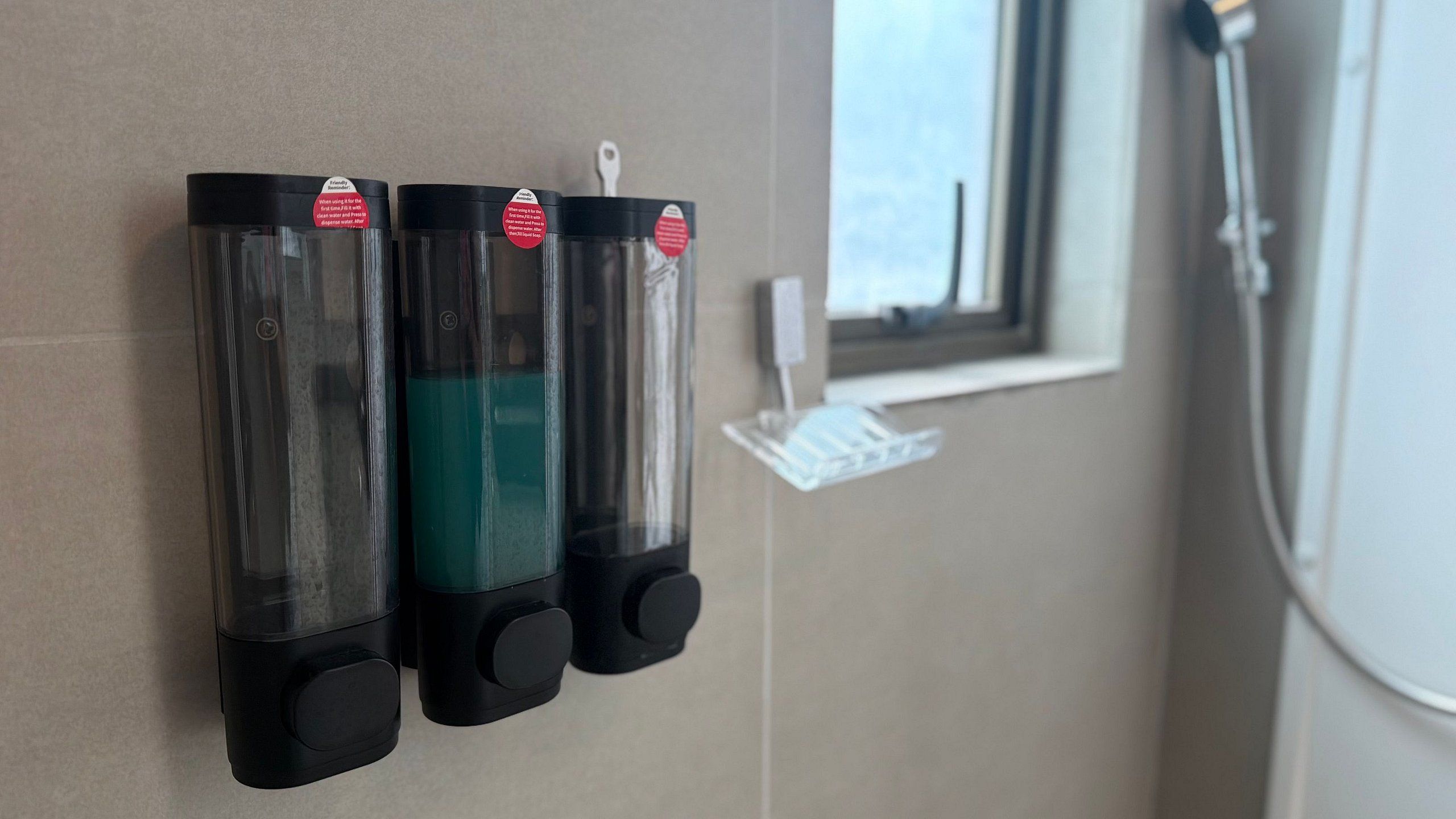 Three black dispensers with red labels on a beige wall, a window with a view, and a white object on a stand.
