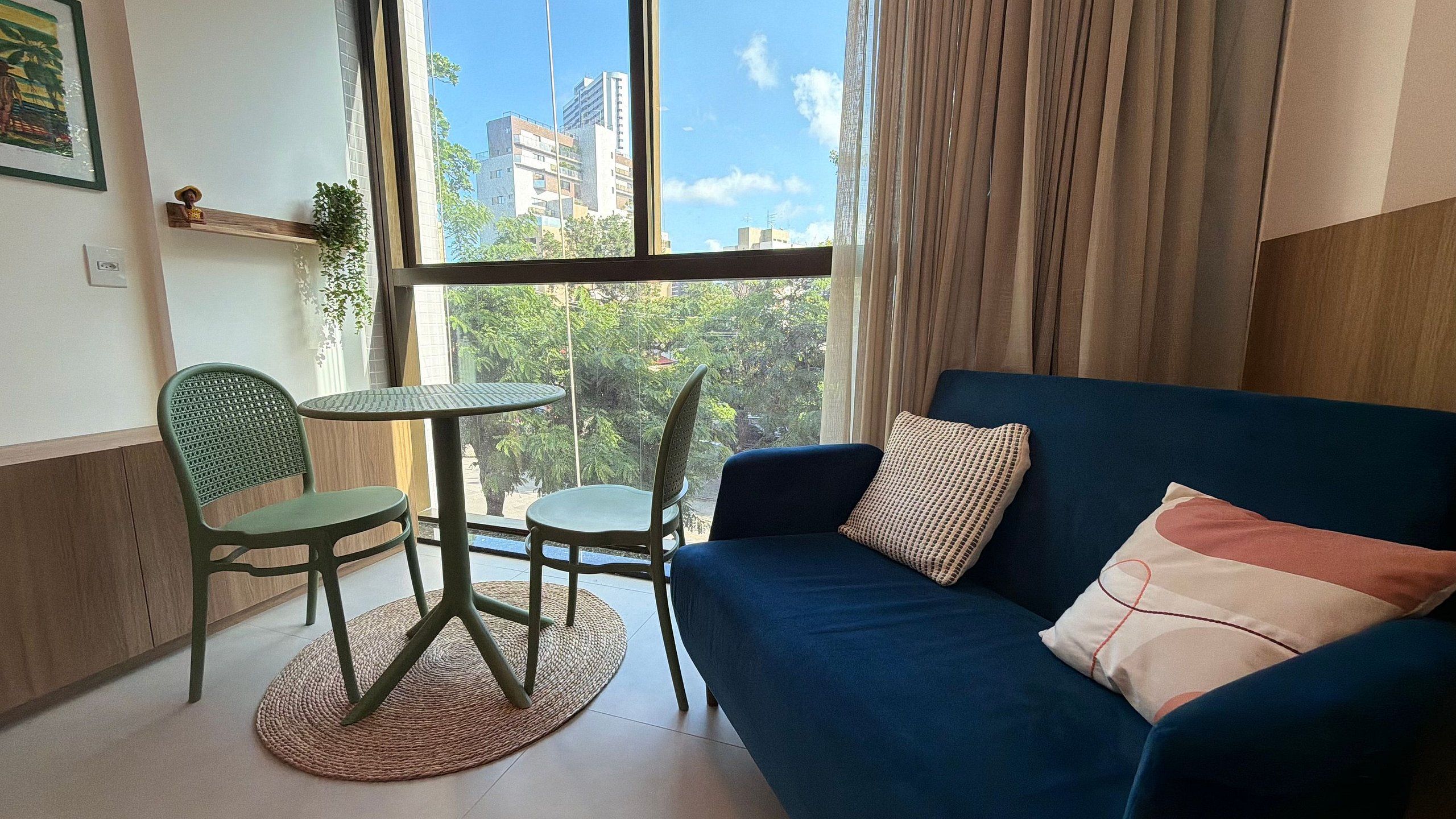 Uma sala de estar aconchegante com um sofá azul, duas poltronas, uma mesa redonda pequena e vista para edifícios através de grandes janelas.