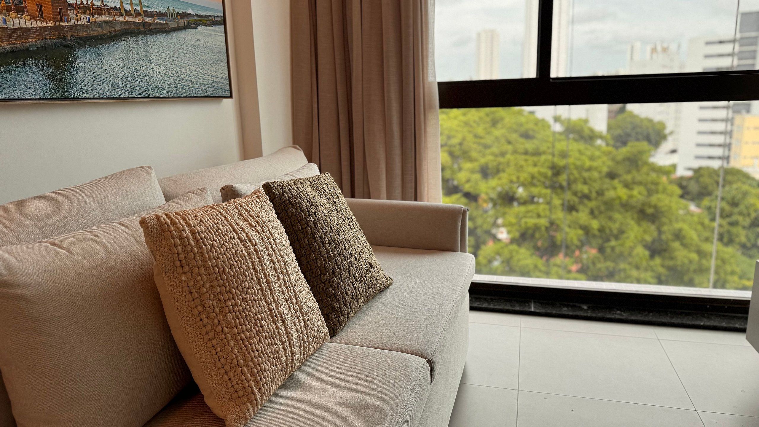 Ein modernes Wohnzimmer mit einer Couch, dekorativen Kissen, einem großen Fenster mit Stadtblick und einem Gemälde an der Wand.