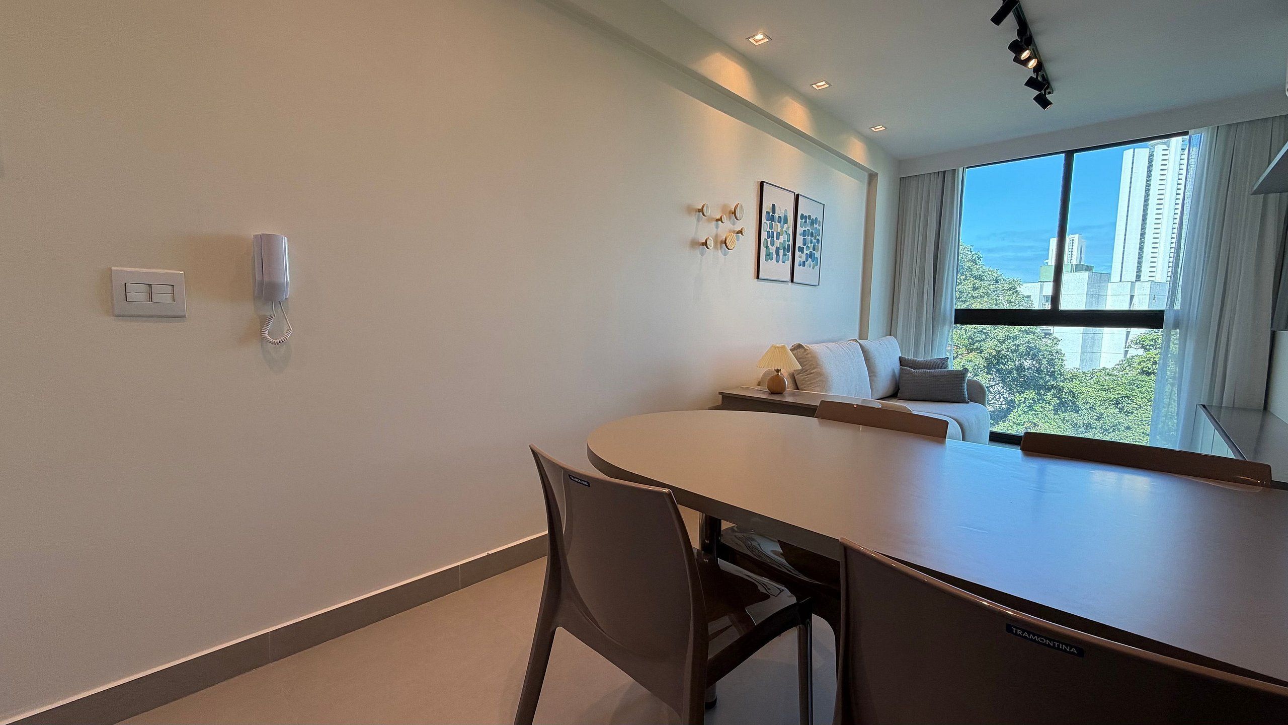 A modern hotel room with a dining table, chairs, a sofa, a lamp, and a view of the cityscape.
