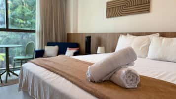 A hotel room with a neatly made bed, a view of greenery outside, and a dining area with a table and chairs.