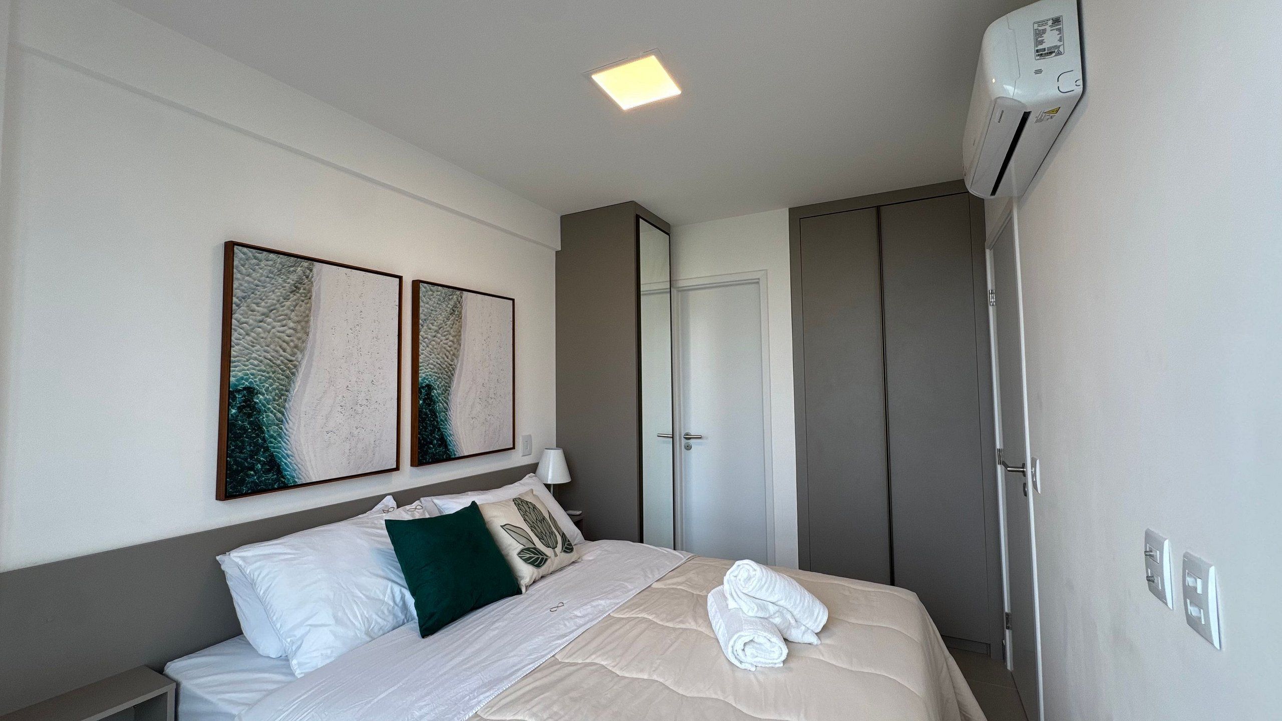Quarto de hotel com cama, duas obras de arte penduradas na parede, abajur, guarda-roupa e ar condicionado.