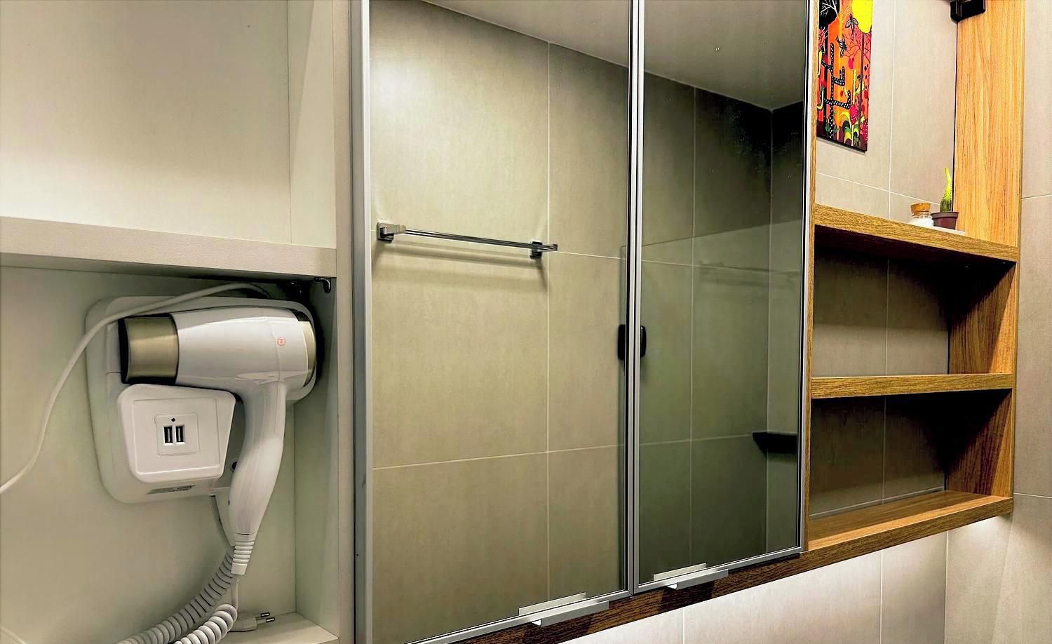 A bathroom with a hairdryer, a glass shower enclosure, and a wooden shelf.