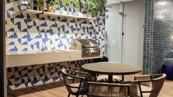 Un patio al aire libre con una mesa y sillas de madera redondas, una parrilla empotrada y un frente de cocina con un diseño geométrico de azulejos.
