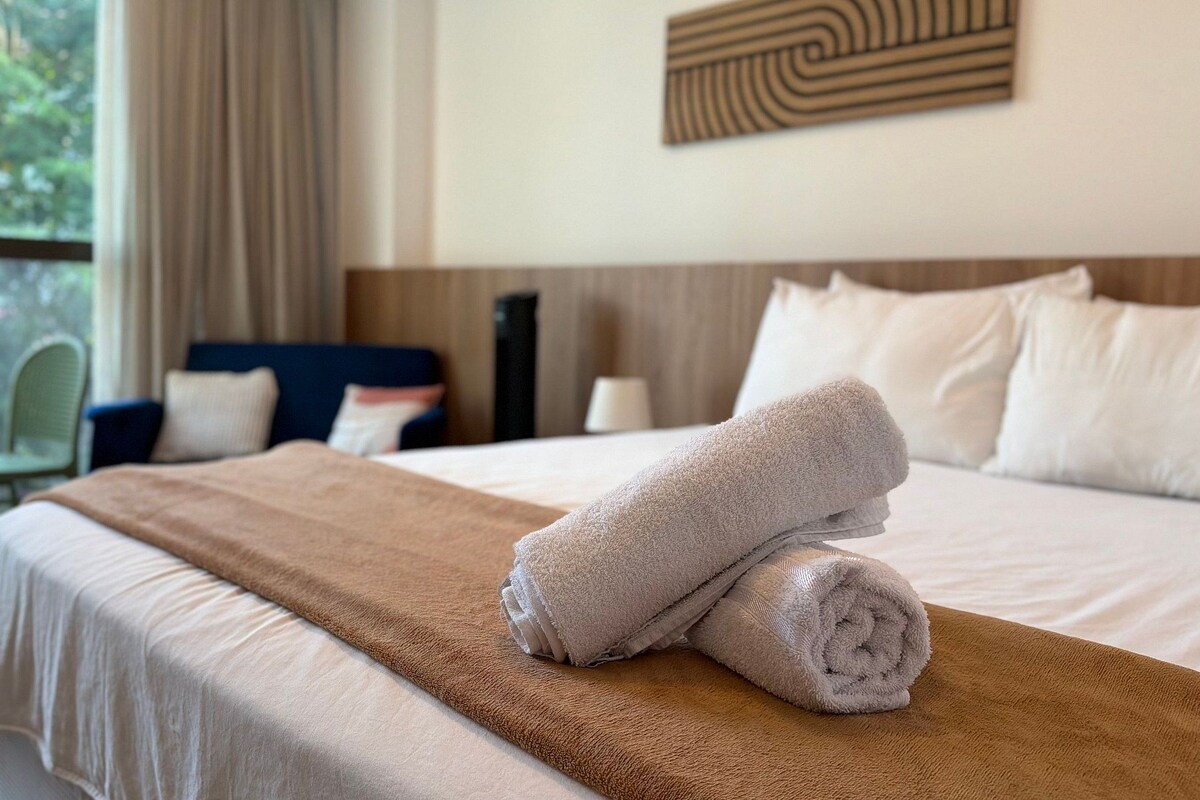 Een hotelkamer met een net opgemaakt bed, uitzicht op groen en een eethoek met een tafel en stoelen.