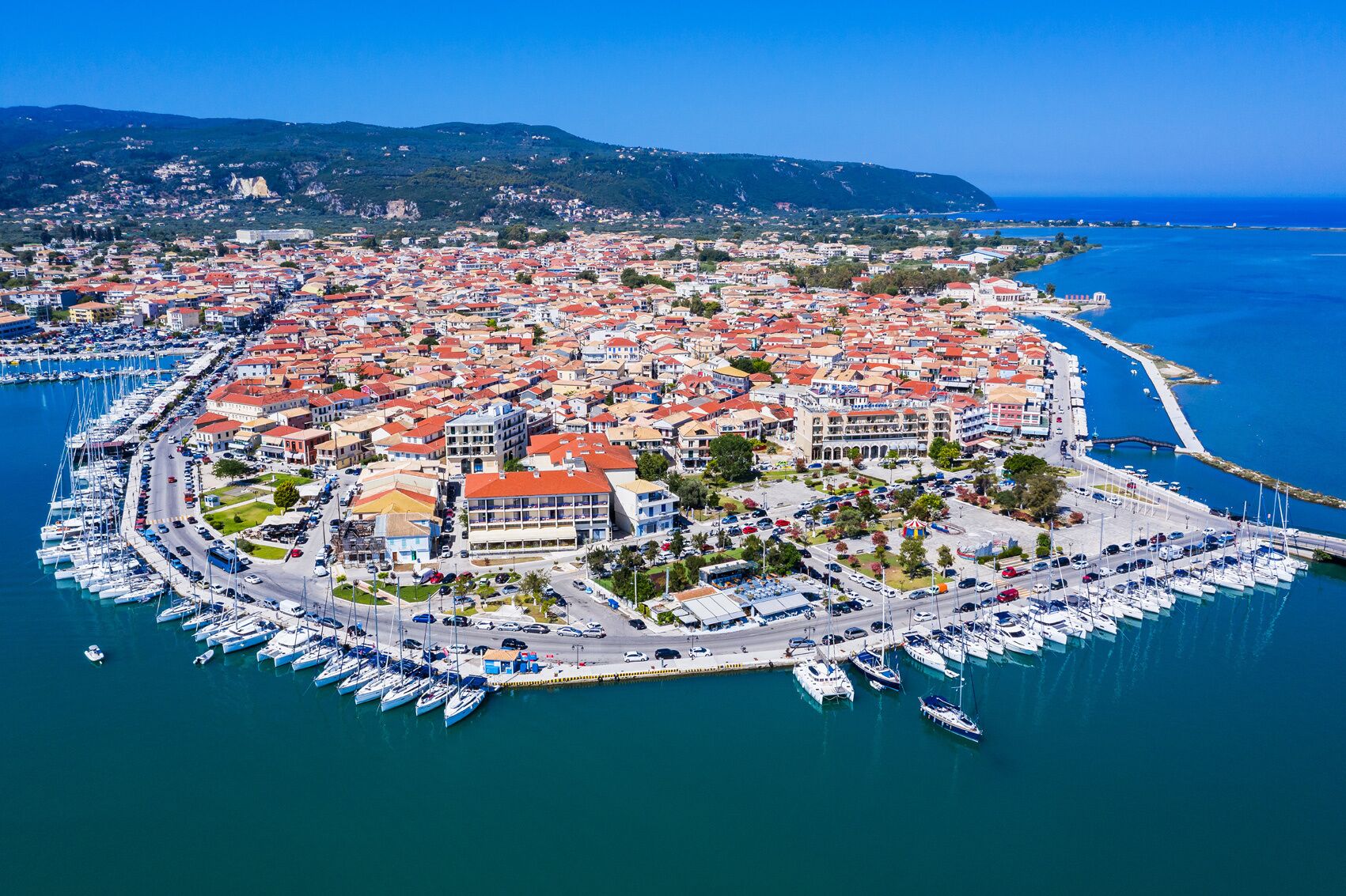 An aerial view of a coastal town with a marina, red-roofed buildings, and a clear blue sea.