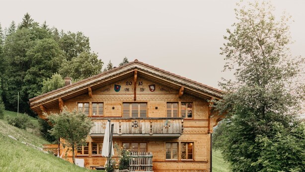 Eine zweistöckige Holzhütte mit Balkon, gelegen an einem Hang inmitten von Grün.